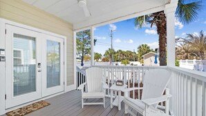 A covered porch with white chairs and a table, a white door with glass panels, and a view of palm trees and houses.