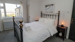 Een net opgemaakt bed met witte lakens, een nachtkastje met een lamp en een raam met gordijnen.