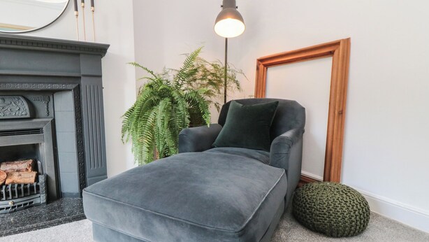 Een knusse woonkamer met een grijze bank, een spiegel met houten lijst, een groene plant en een open haard met hout.