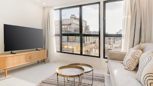 A modern living room with a large flat-screen TV on a wooden console, a sofa with patterned cushions, and a large window offering a city view. - Ototo Luxury Living (Thessaloniki)