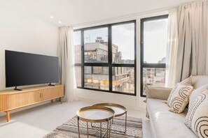 Ein modernes Wohnzimmer mit einem großen Flachbildfernseher auf einer Holzkonsole, einem Sofa mit gemusterten Kissen und einem großen Fenster mit Blick auf die Stadt.