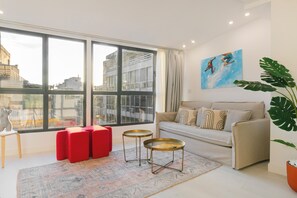 Un salon moderne avec une grande fenêtre, un canapé, des poufs rouges, deux petites tables rondes et une grande plante.