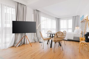 Une chambre d’hôtel moderne équipée d’un grand téléviseur à écran plat monté sur un trépied, d’une table de salle à manger ronde avec des chaises en osier, d’un canapé et d’un luminaire suspendu.