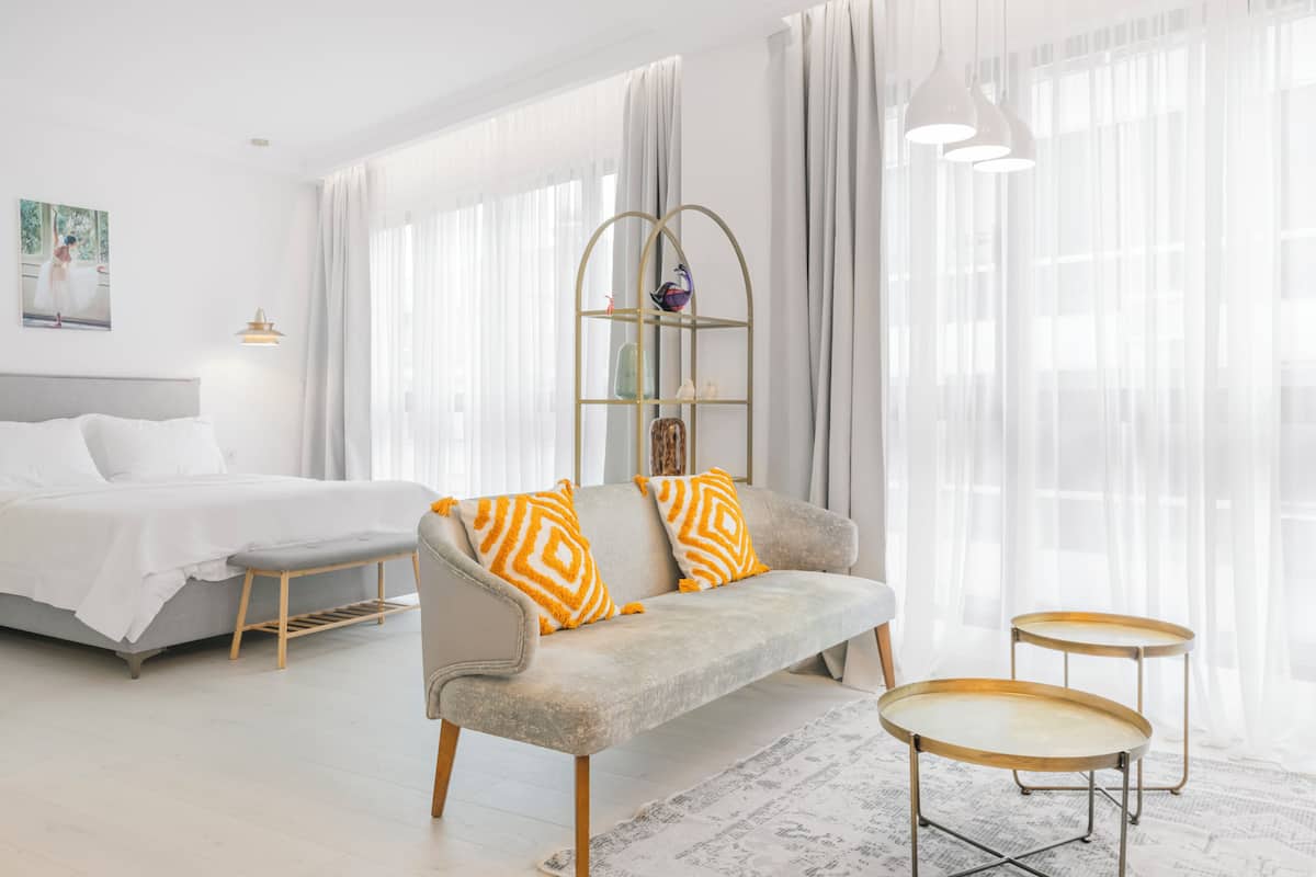 Ein modernes Hotelzimmer mit grauem Sofa, einem Bett, zwei kleinen Tischen und transparenten Vorhängen.