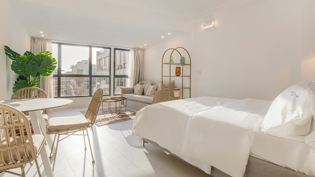 A modern hotel room with a bed, a sofa, a dining table with chairs, a large window, and a potted plant. - Ototo Luxury Living (Thessaloniki)