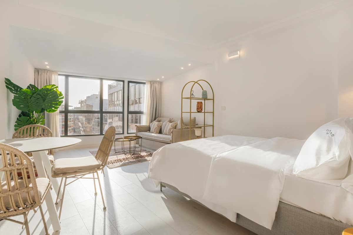 Ein modernes Hotelzimmer mit Bett, einer Couch, einem Esstisch mit Stühlen, einem großen Fenster und einer Zimmerpflanze.
