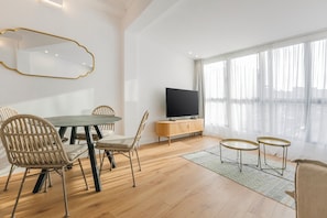 Una sala de estar moderna con una mesa de comedor, sillas, un televisor y un ventanal con cortinas traslúcidas.