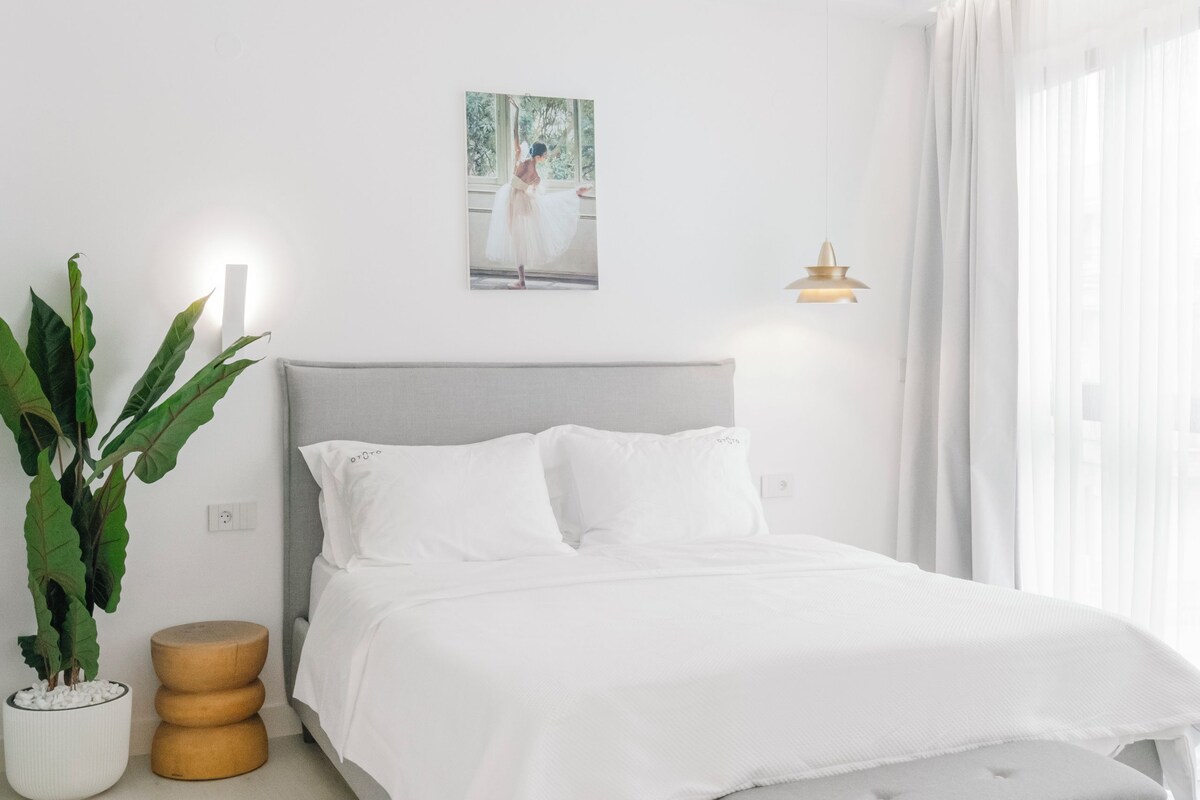 Une chambre moderne avec un lit gris, du linge de lit blanc, un banc gris, une plante en pot et un luminaire fixé au mur.
