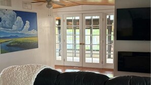 A living room with a black leather couch, a painting of a landscape, a ceiling fan, and a television on the wall. - The Bamboo house on the lake (Hernando)