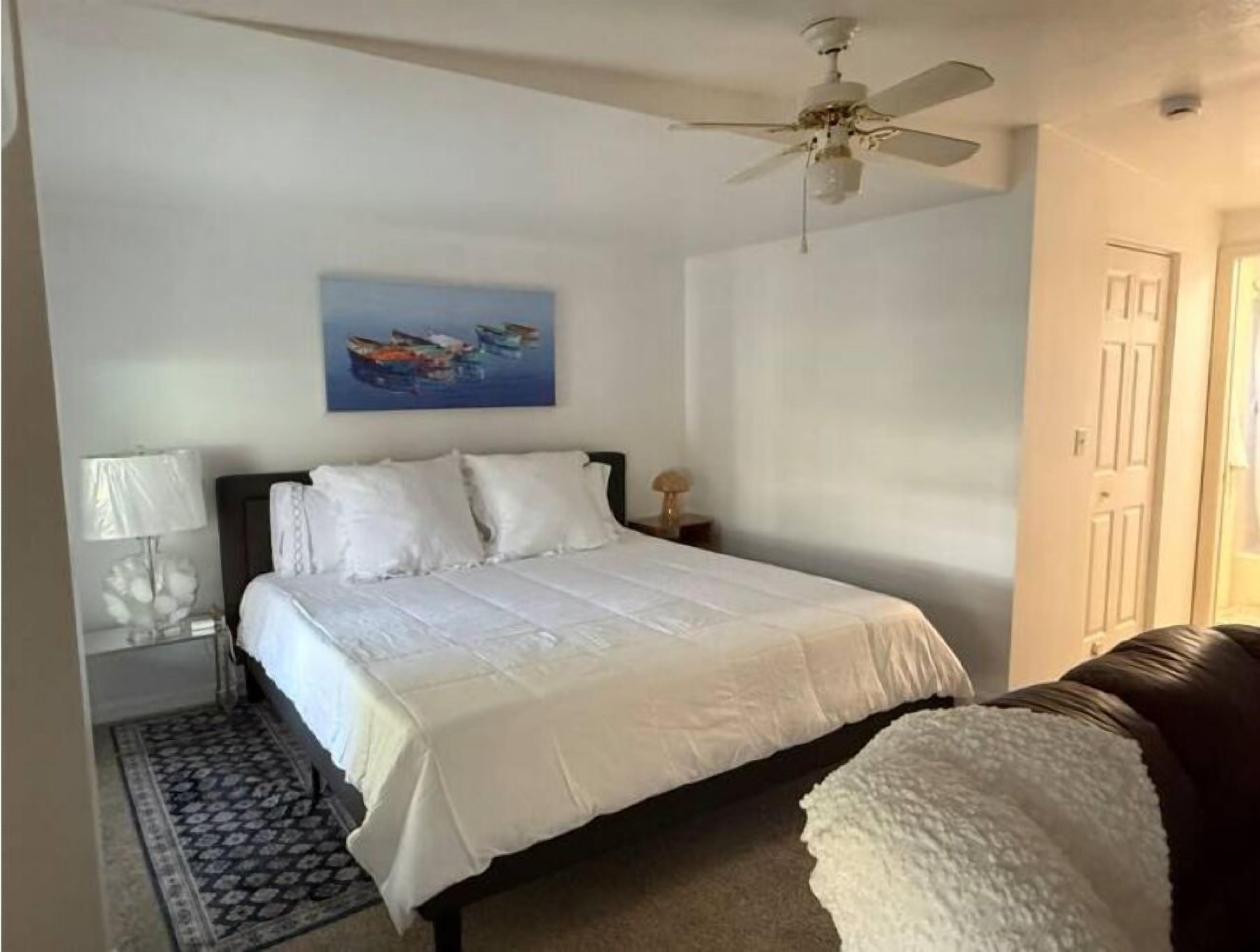 A bedroom with a bed, a ceiling fan, a nightstand, a lamp, and a painting on the wall.