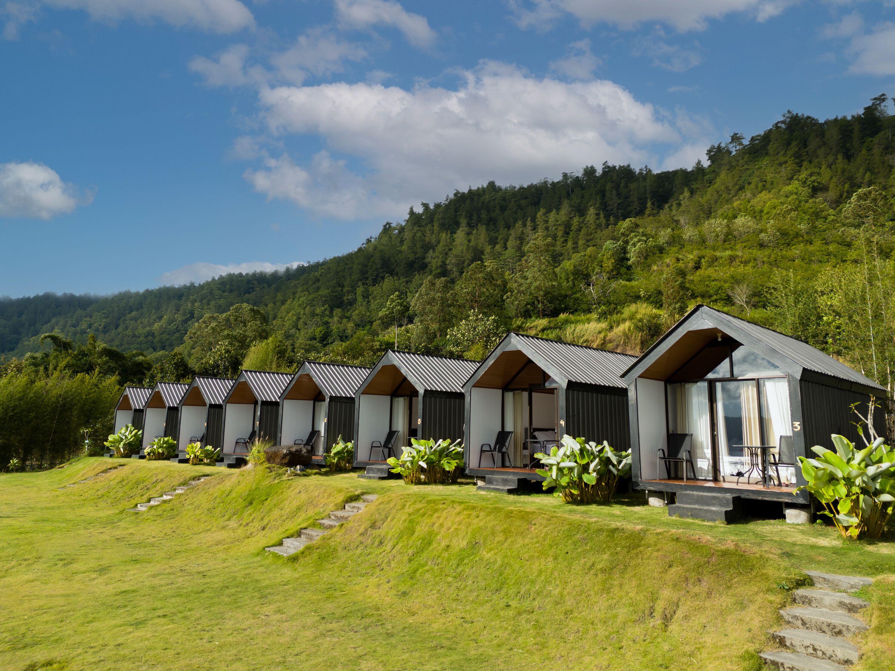A row of cabins with a grassy slope leading up to them, set against a backdrop of a forested hill.