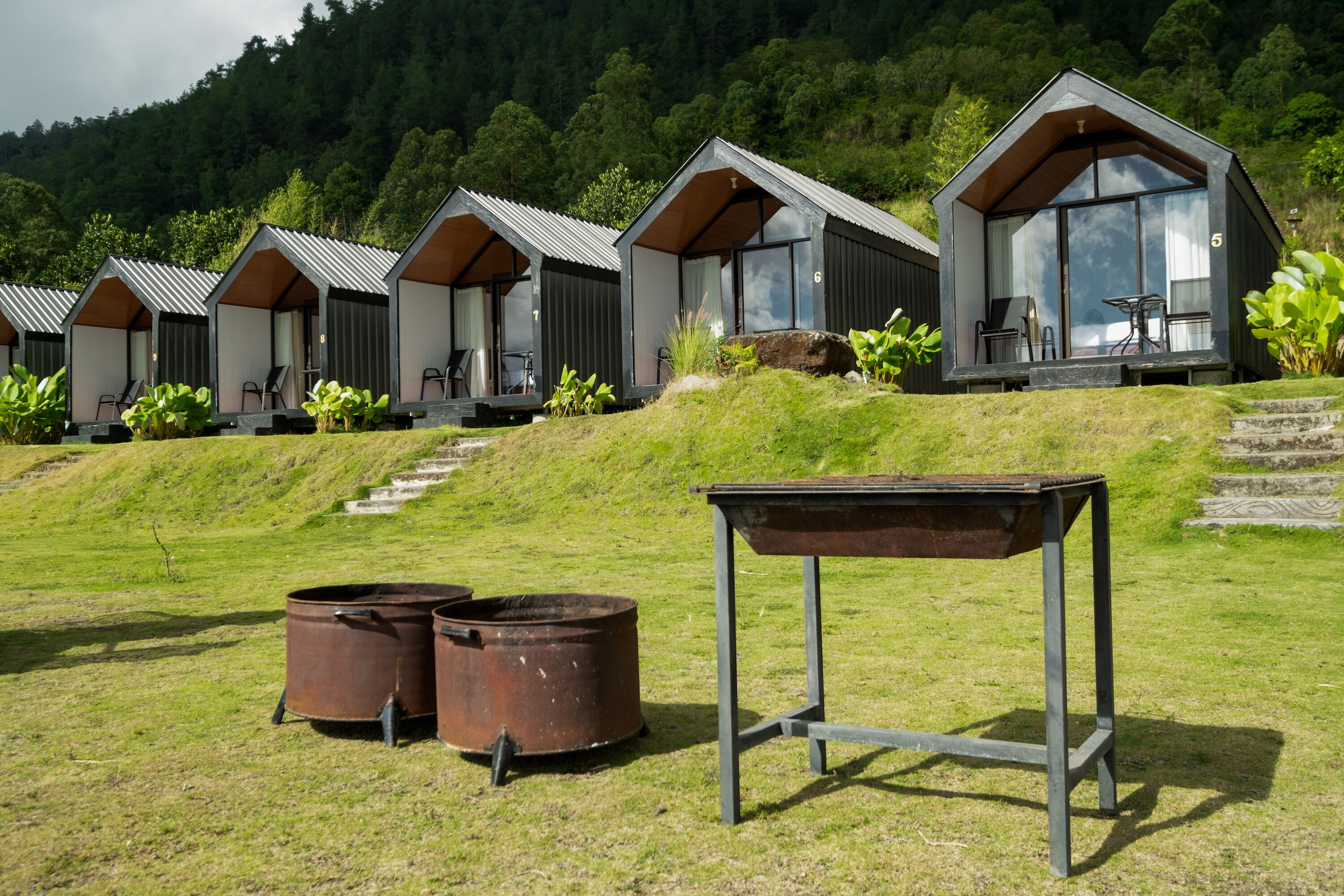 A row of modern, single-story cabins with large windows, set on a grassy hill with a forested backdrop.