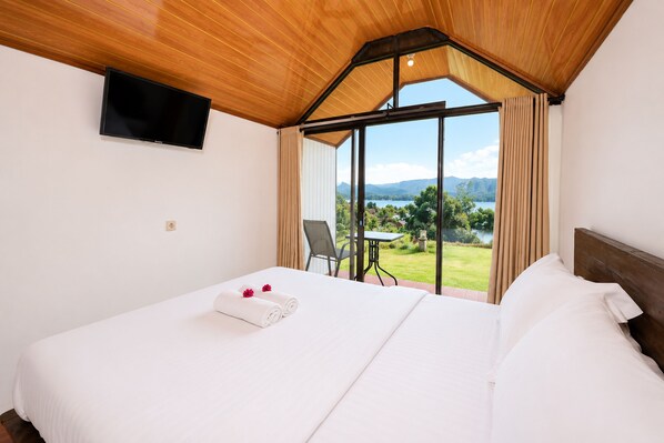 A hotel room with a bed, a TV mounted on the wall, a window with curtains, and a view of a lake and mountains. - Royal Indah Cabin (Kintamani)