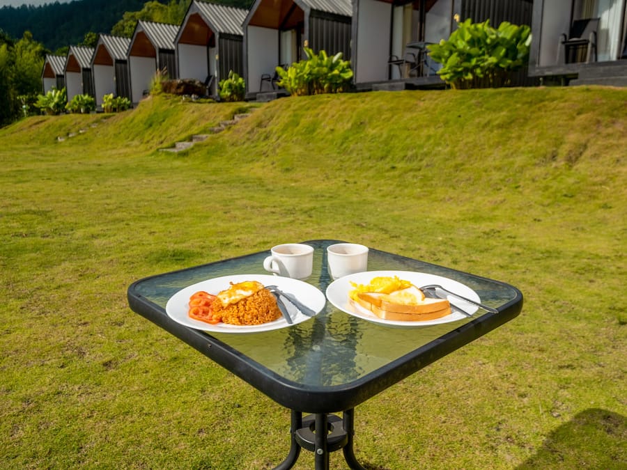 Una mesa con comida y dos tazas, sobre un césped con edificios al fondo.