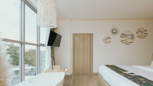 A modern bedroom with a large window, a bathtub, a bed with a patterned bedspread, and wall art. - Parima Hotel (Trang)