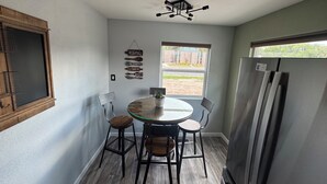 Una cocina con una mesa redonda de vidrio, dos sillas, un refrigerador y una ventana con vistas a un edificio y vegetación.