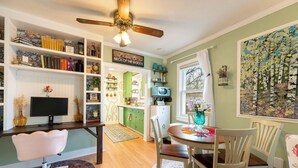 A cozy room with a wooden desk, a bookshelf, a dining area with chairs, a ceiling fan, and a large window with curtains.