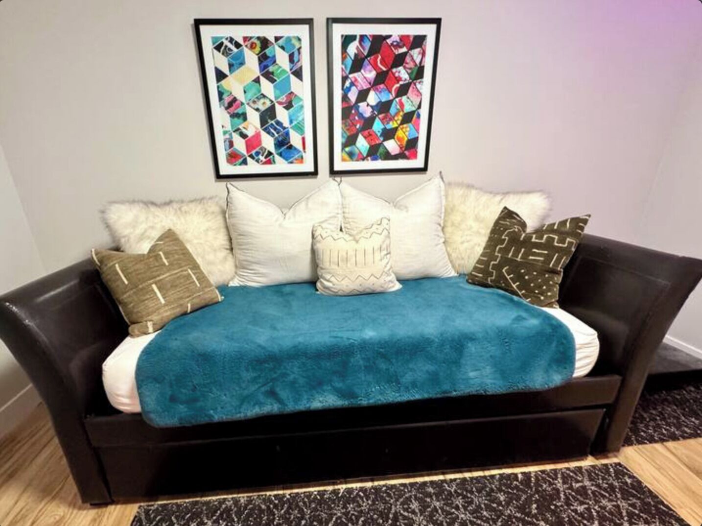 Un sofá negro con una manta color aguamarina, almohadas blancas con diseños, y dos cuadros enmarcados arriba.