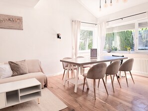 A modern living room with a sofa, a dining table with chairs, a white coffee table, and a view of greenery through the window. - Holiday Home 'Landhausglück' with Private Terrace, Private Garden and Wi-Fi (Geislingen)