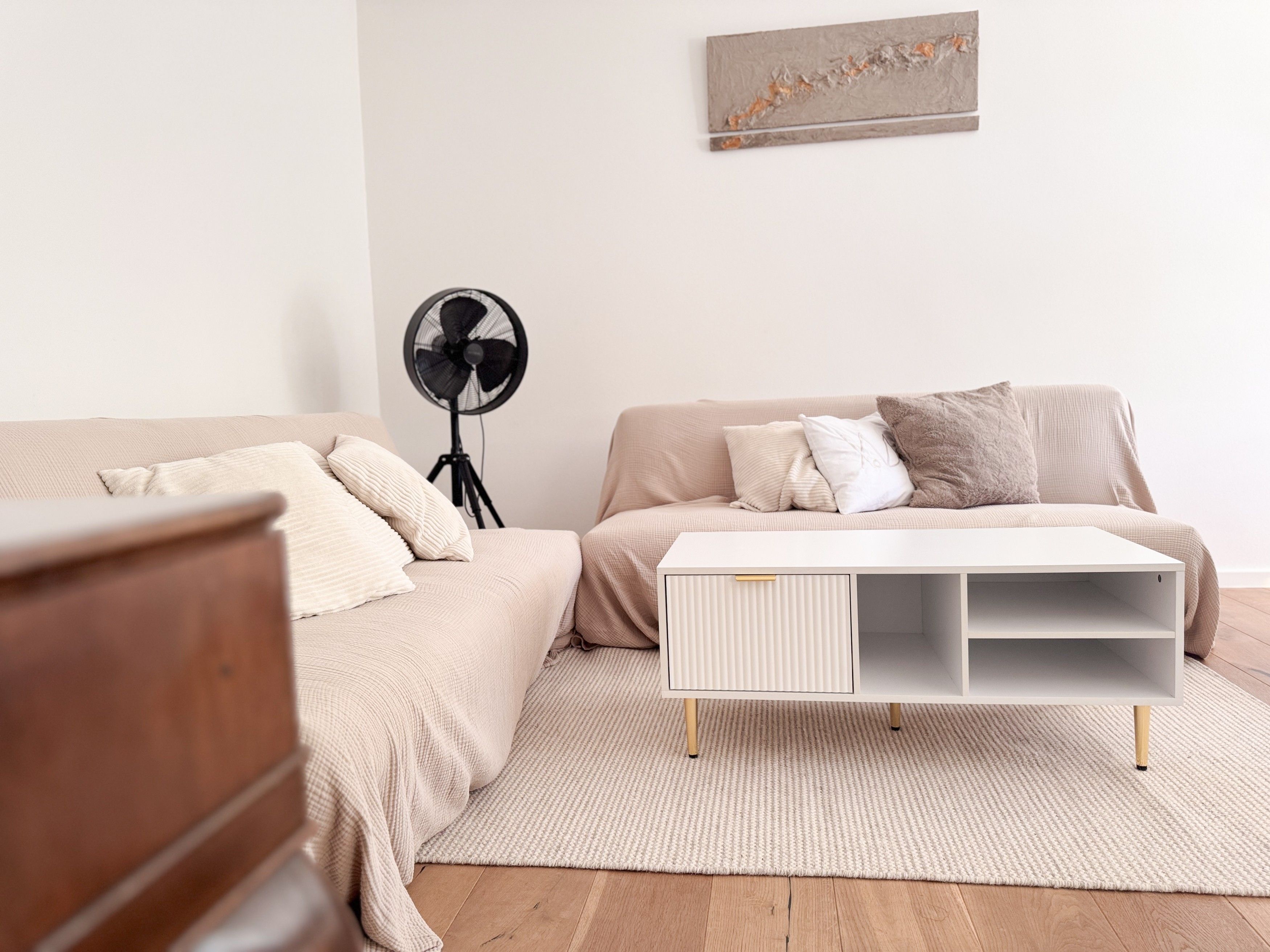 A bedroom with a bed, a white side table, a floor fan, and a wall-mounted artwork.