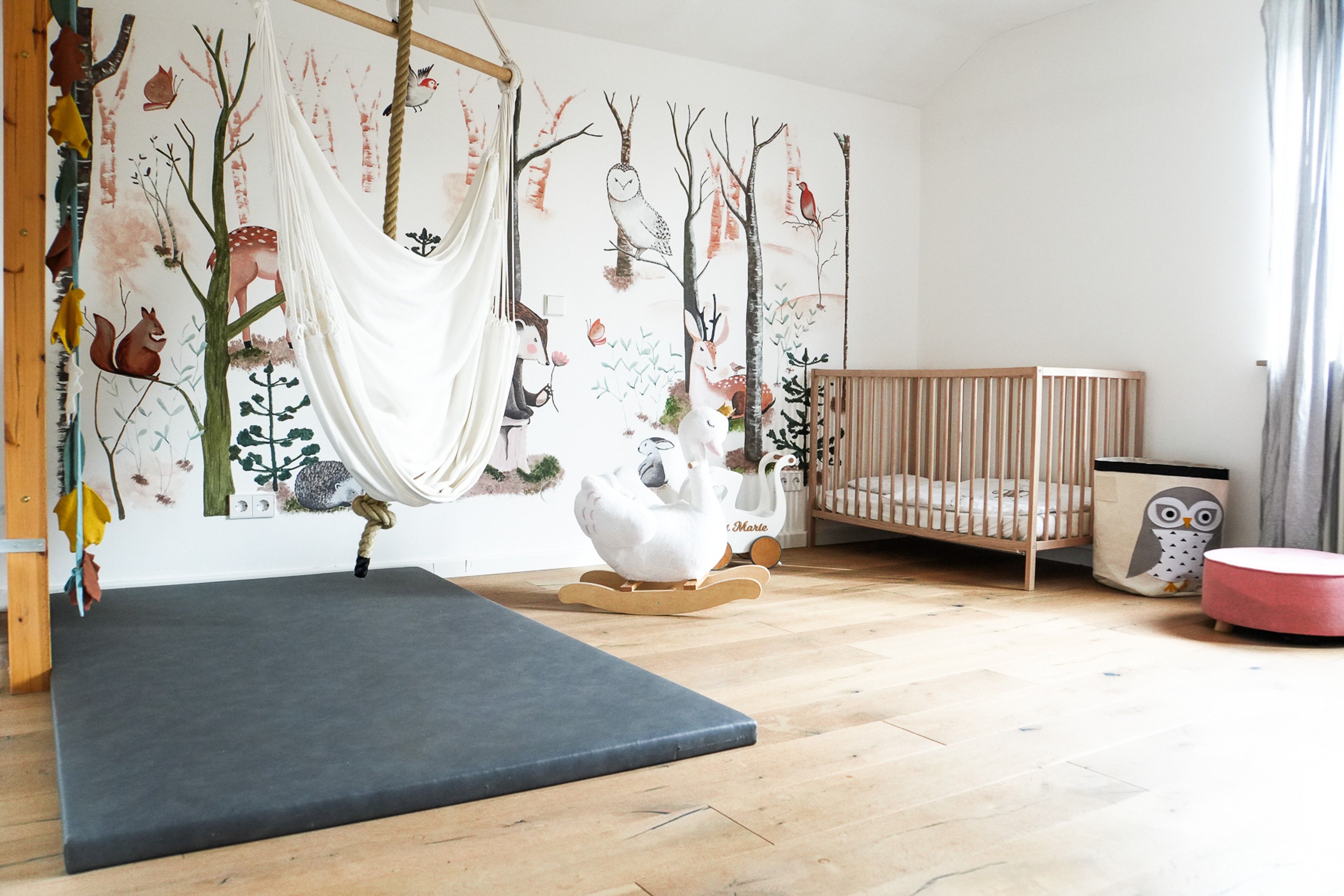 Ein Zimmer mit Hängekorbsessel, Babybett und Schaukelpferd, sowie einer Wandmalerei mit Waldbewohnern.