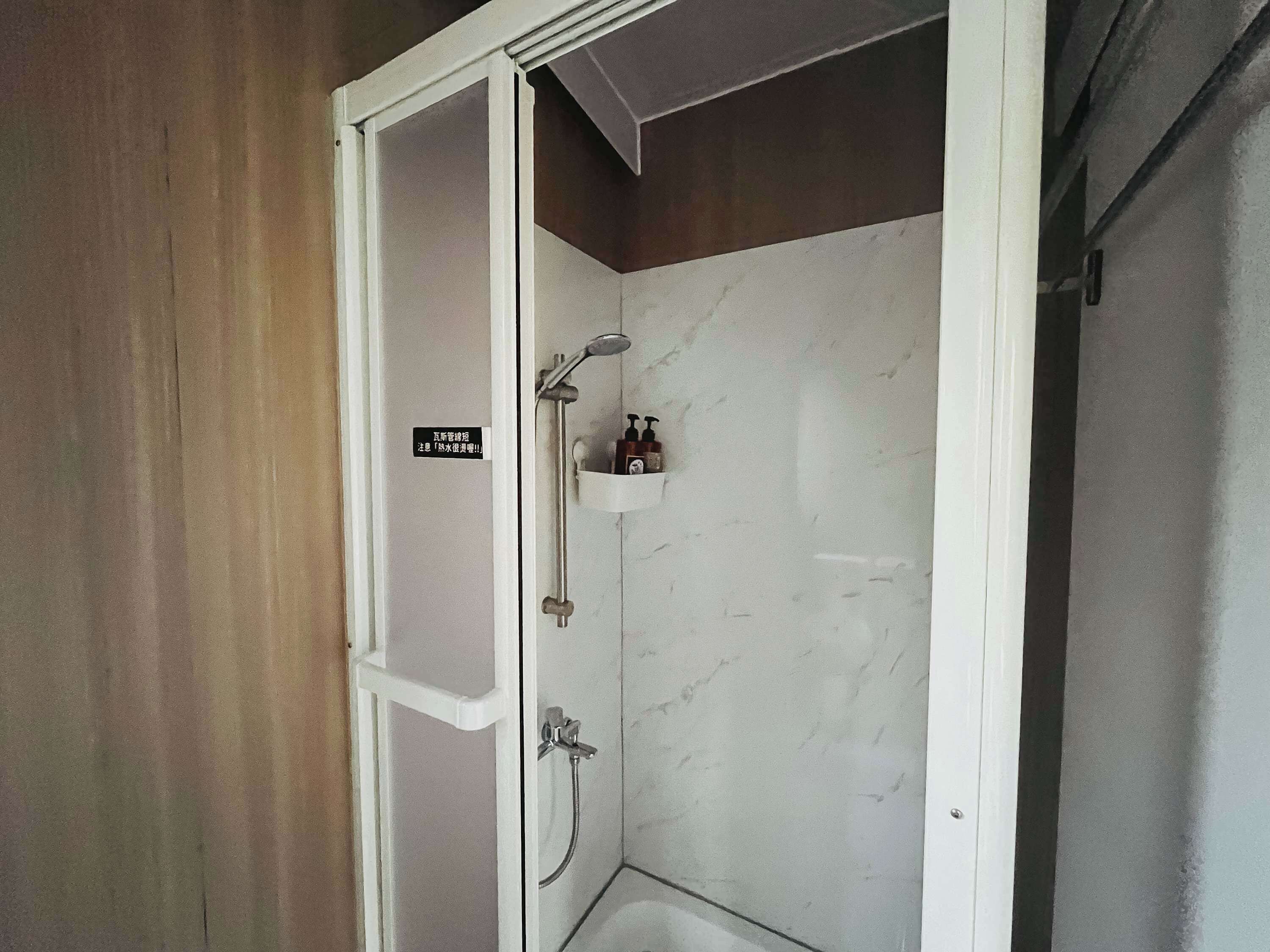 A shower with a handheld showerhead and a wall-mounted faucet, a small shelf with toiletries, and a sign on the door.
