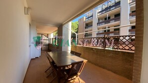 Une terrasse extérieure couverte, dotée d’une table et de chaises, d’un mur en briques et offrant une vue sur un immeuble à plusieurs étages avec des balcons.