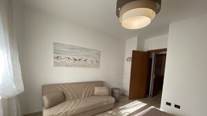 Une chambre avec un canapé beige, un lit, une petite table ronde et un tableau au mur.