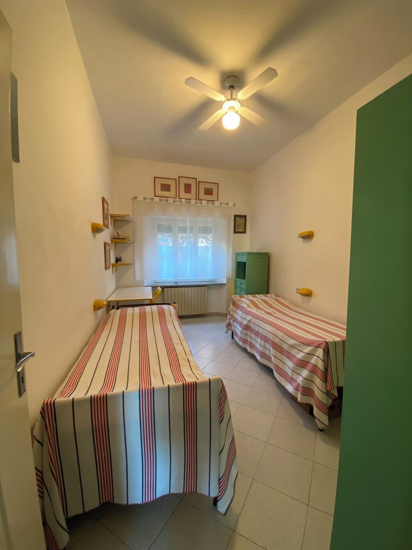 A small, single-room accommodation with two single beds, a ceiling fan, a window with a curtain, and a radiator.