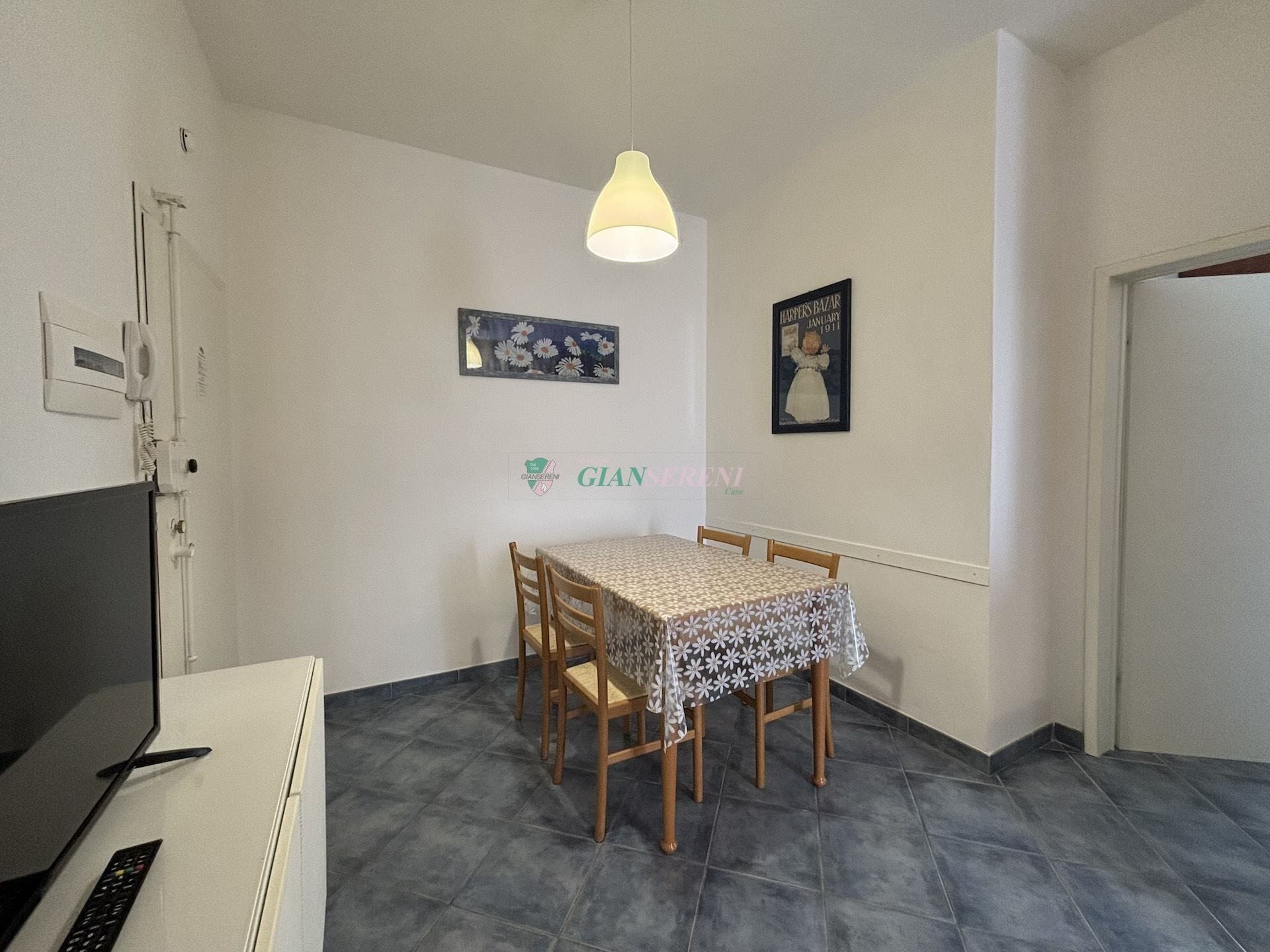 A dining area with a table covered by a floral tablecloth, wooden chairs, a ceiling light, and a refrigerator.