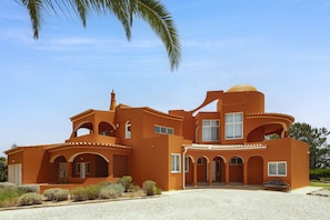 A large, orange building with multiple arches and a dome, surrounded by a paved driveway and greenery.
