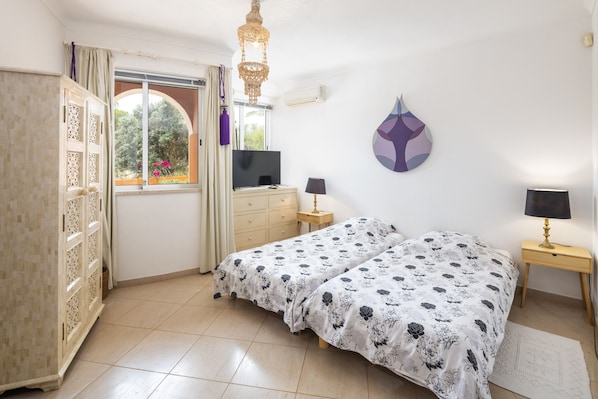 A bedroom with two beds, a TV, a chandelier, and a window with curtains.