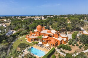 En stor, orange villa med en swimmingpool, omgiven av frodig grönska och med utsikt över havet.