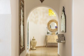 A hallway with a decorative mirror, a round mirror, a vase, and a cabinet with decorative items.