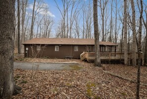 A cabin with a wooden deck and a small porch, surrounded by trees and a leaf-covered ground.