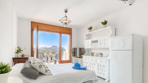 Une chambre à coucher comprenant un lit, une commode, un téléviseur et un balcon avec vue sur les immeubles.