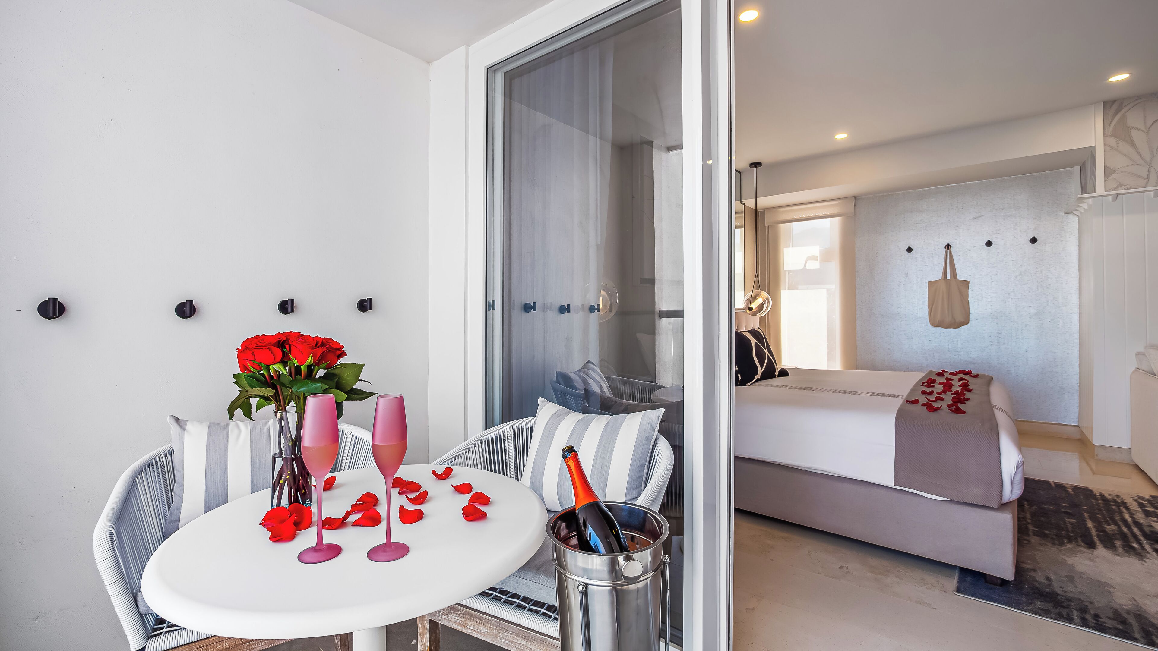 A modern room with a glass partition, a bed with a white bedspread, a bedside table with a bottle and glasses, and a vase with red roses.