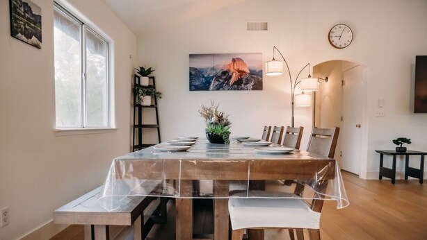 A dining area with a wooden table, chairs, and a clear plastic tablecloth. There is a potted plant on the table, a clock on the wall, and a framed picture above the table.