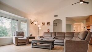 A modern living room with a sofa, armchair, and coffee table. There are large windows, a ceiling fan, and wall-mounted lamps.