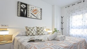 Un dormitorio ordenado con cama, almohadas, lámpara de noche, un libro y una ventana con cortinas.