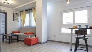 Una sala moderna con sofá, reposapiés y una mesita. Hay una ventana con cortinas, una zona de cocina con un taburete de bar y un perchero.