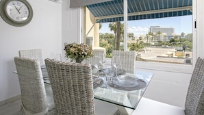 Un comedor con sillas de mimbre, una mesa de vidrio y vistas a palmeras y edificios.