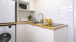Een kleine keuken met een wasmachine, een broodrooster, een spoelbak, en een klein aanrecht met een potplant en fruitschaal.