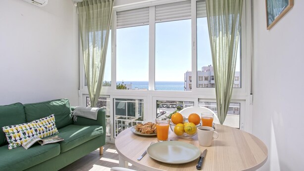 Een kamer met een groene bank, een eettafel met ontbijtartikelen en uitzicht op zee door grote ramen.