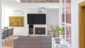 Ein modernes Wohnzimmer mit grauem Sofa, einem an der Wand montierten Flachbildfernseher, einem Kamin und einem Essbereich mit Stühlen und Tisch.