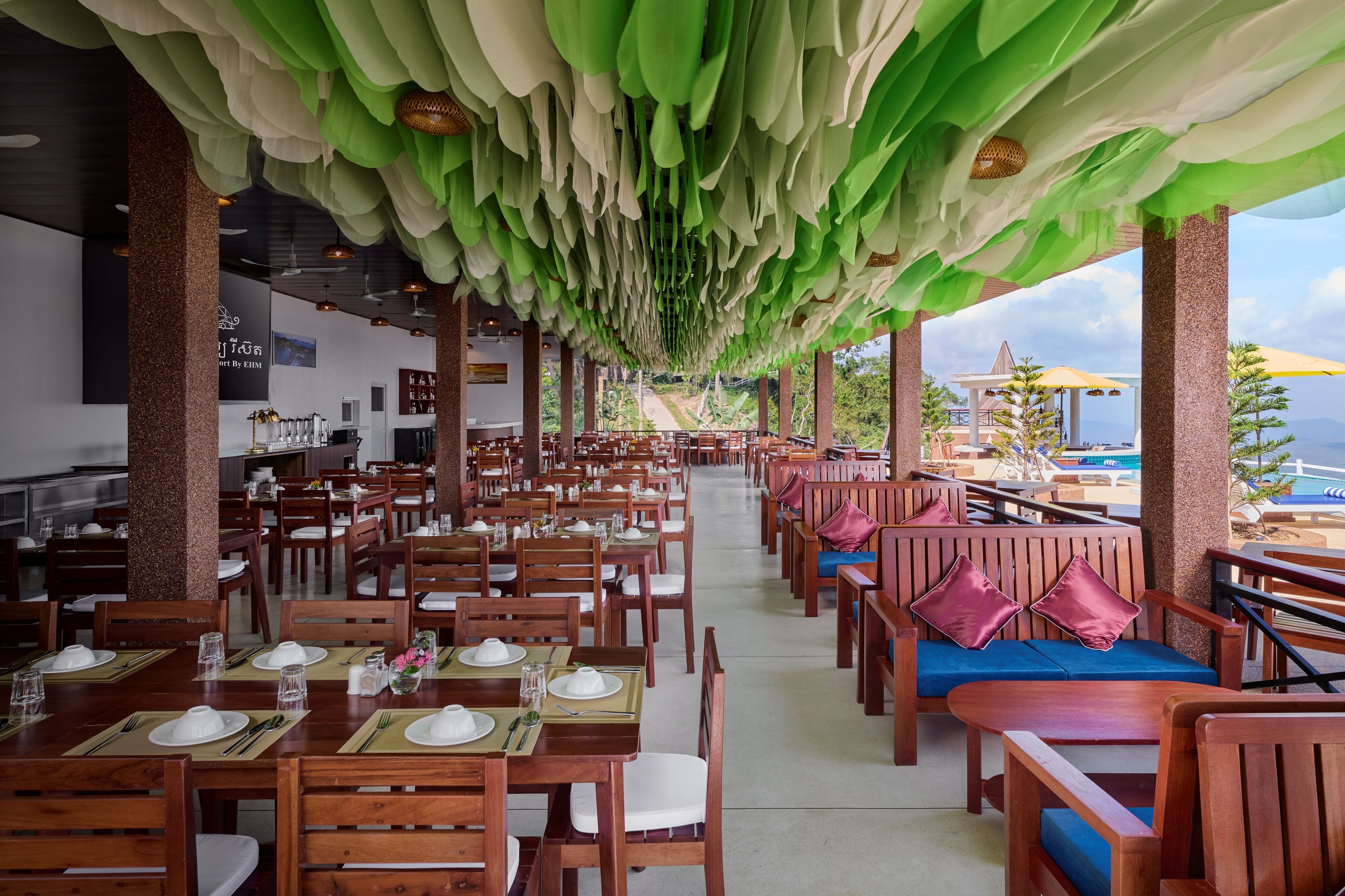 Um restaurante com mesas e cadeiras de madeira, detalhes decorativos verdes no teto e vista para o mar.