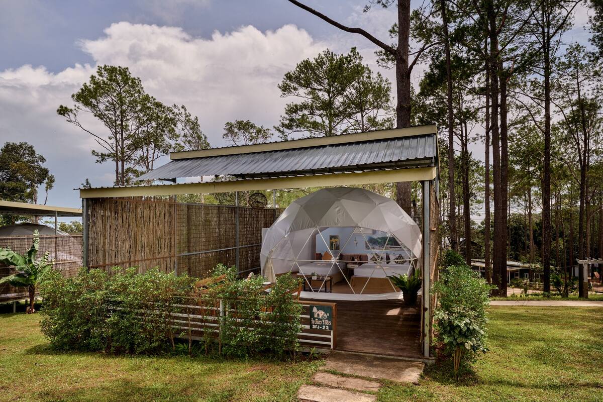 A geodesic dome structure with a wooden deck and a metal roof, surrounded by trees and a grassy area.
