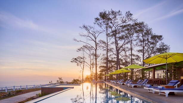 A poolside area with lounge chairs and umbrellas, surrounded by trees and overlooking a body of water.