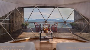 A cozy, open-plan room with a wooden floor, two cushioned chairs, a small table with a fruit basket, and a view of mountains and water through a large window.