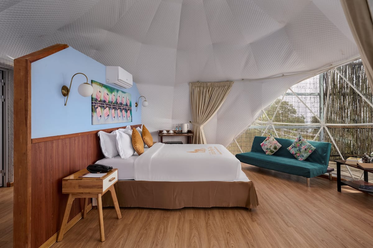A modern hotel room with a large bed, a sofa, a desk, and a large window with a view of trees.